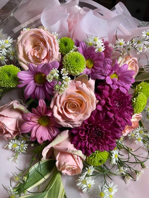 A vibrant floral arrangement featuring soft pink roses with delicate, fully opened petals, complemented by purple daisies with bright yellow centers, and contrasting green button chrysanthemums with r