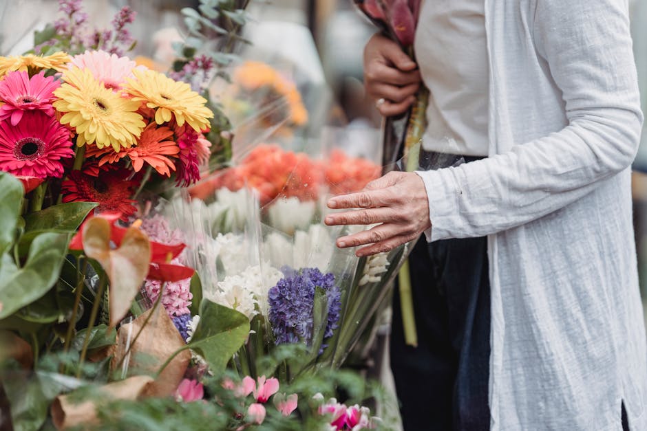 A person selecting vibrant, fresh flower bouquets at an outdoor market, with an array of colorful blooms including pink, yellow, orange, purple, and white daisies, hyacinths, and other flowers arrange