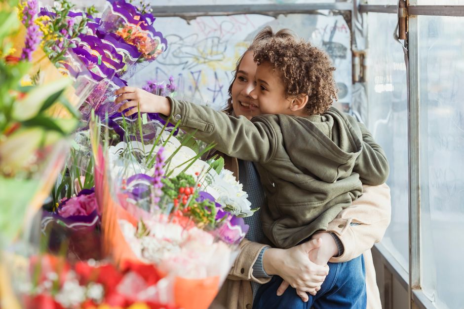 A young child with curly hair and a smiling expression is being held by an adult, reaching out to touch an assortment of colorful flower bouquets on display at a flower shop. The flowers include pink,